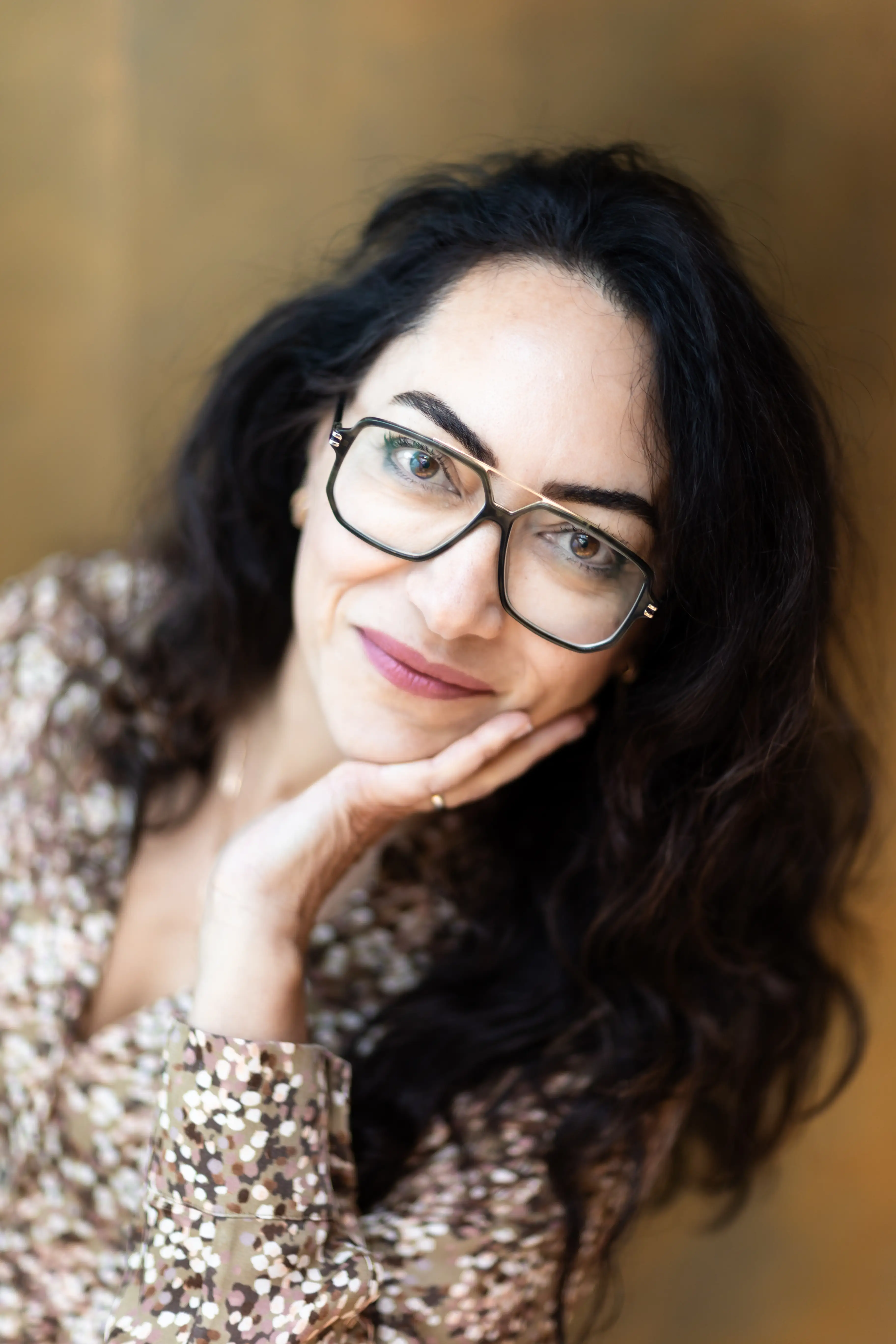 Kobieta z długimi ciemnymi włosami i w okularach uśmiecha się delikatnie, opierając podbródek na dłoni. Ma na sobie wzorzysty top i jest przedstawiona na miękkim, rozmytym tle.
