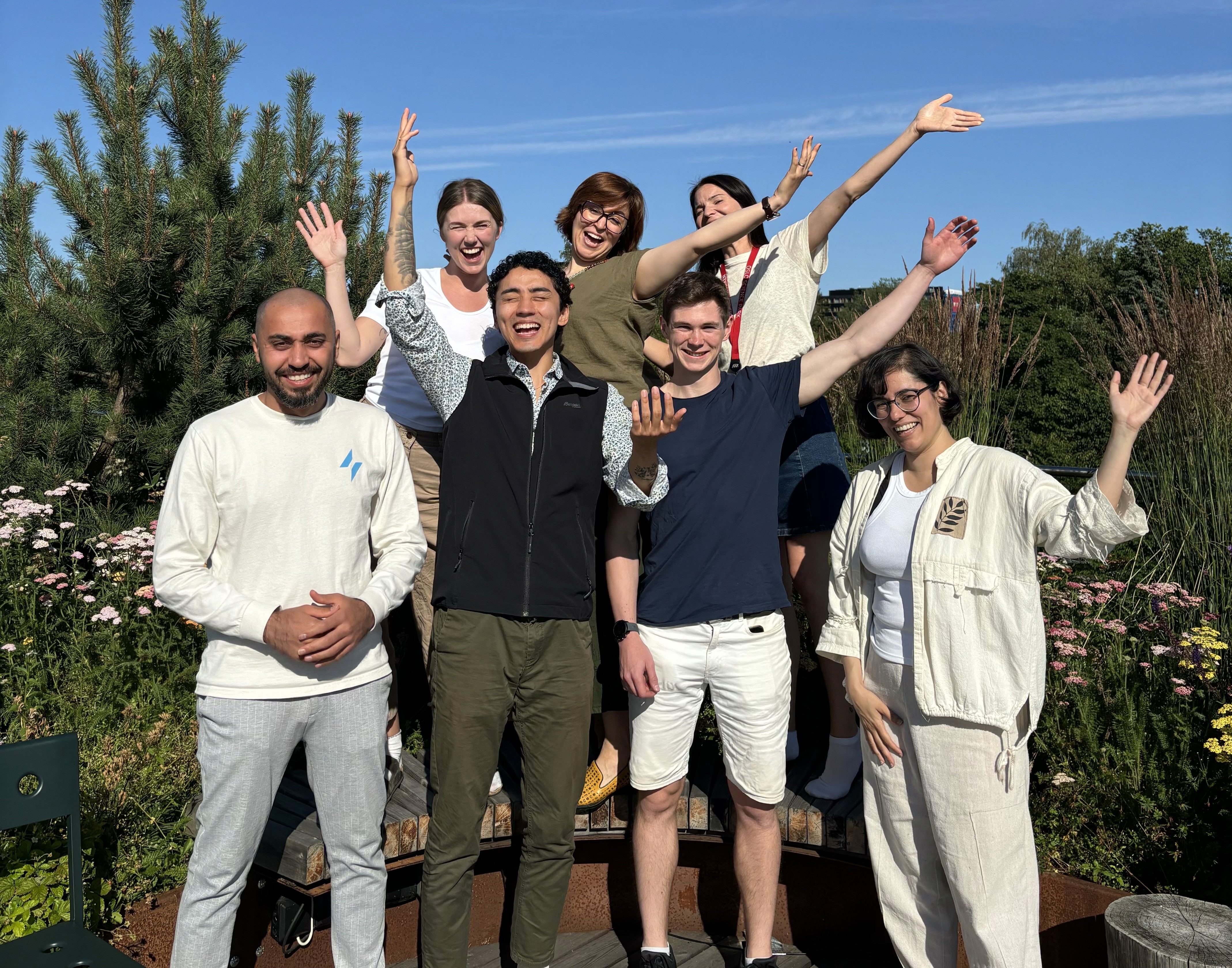 A young person with curly hair smiles at the camera while sitting in front of a laptop against a dark background.