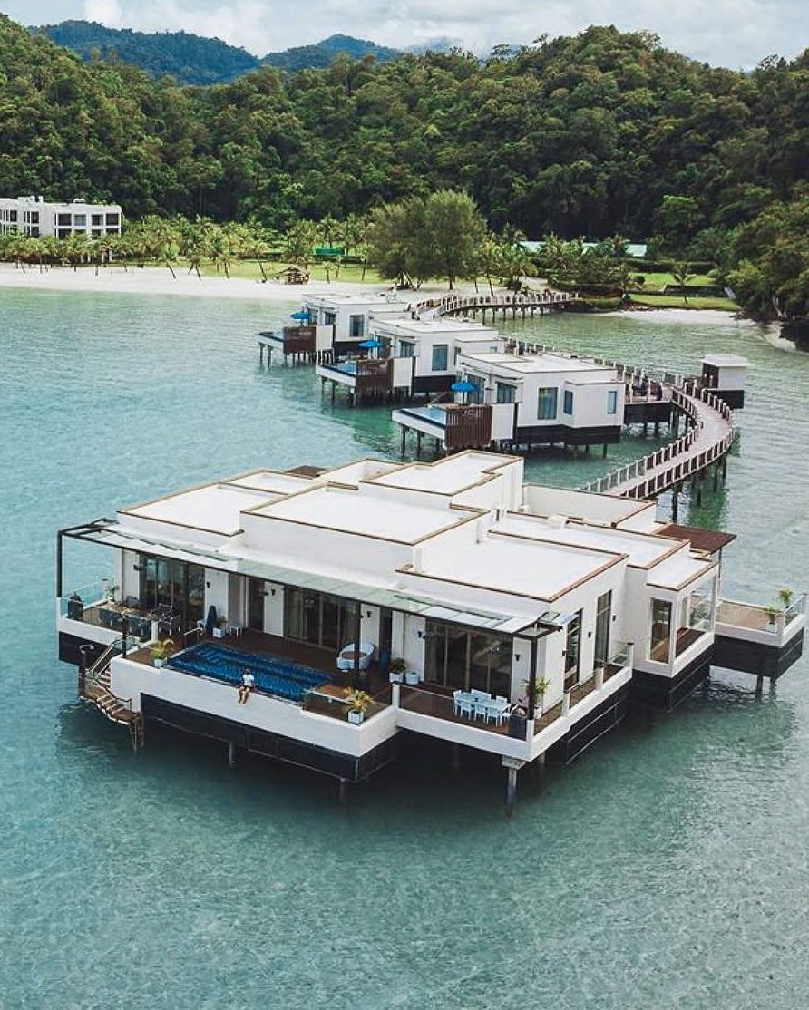 Aerial view of The St. Regis Langkawi resort featuring overwater suites connected by wooden walkways, surrounded by tranquil waters and lush greenery.