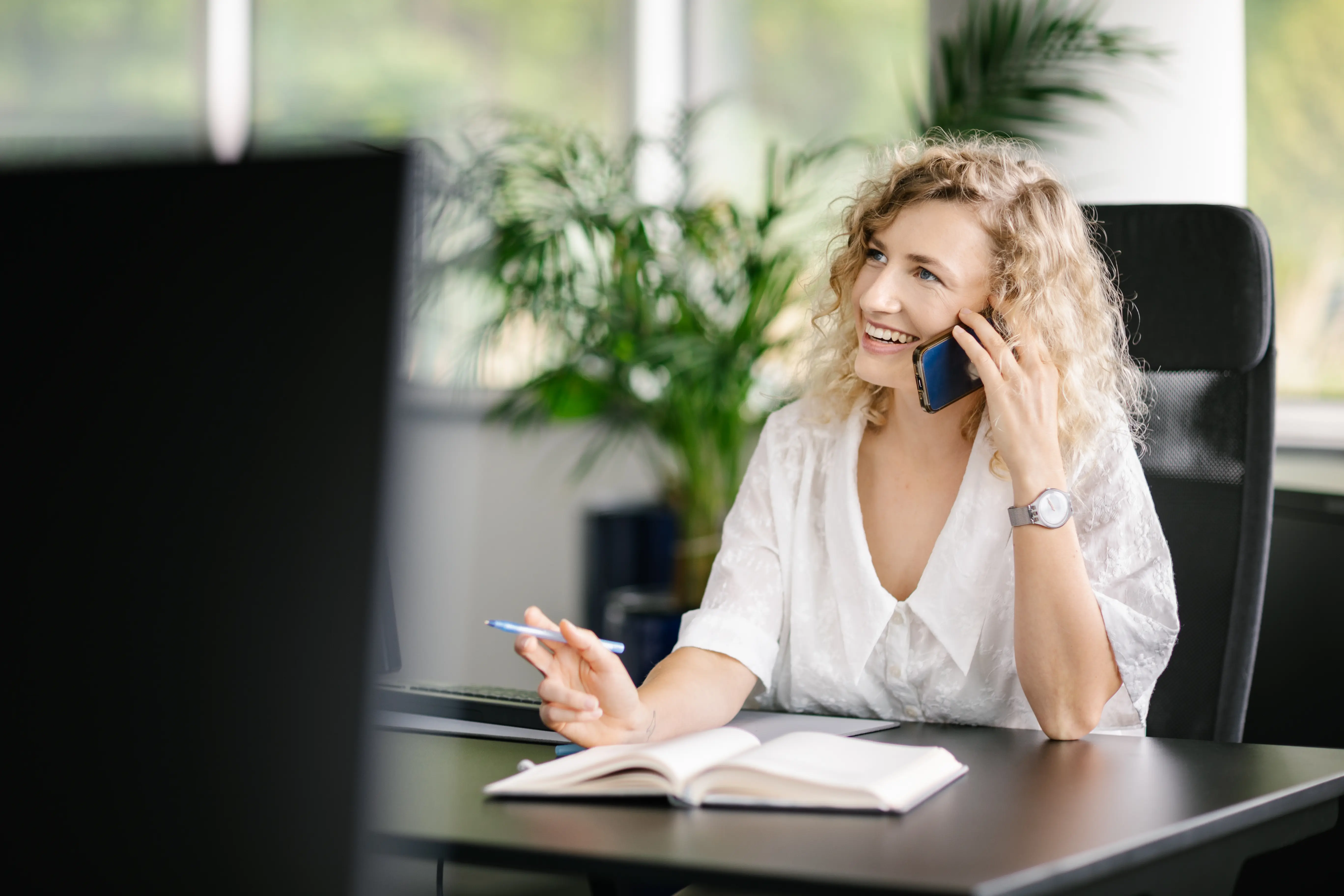 Kobieta o kręconych blond włosach siedzi przy biurku, uśmiechając się i rozmawiając przez smartfona. Trzyma długopis nad otwartym notatnikiem. W tle widać rośliny i duże okna.