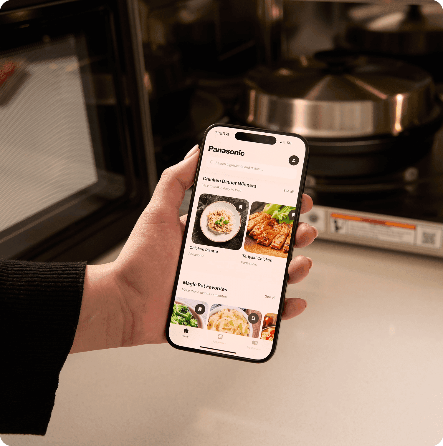 Hand holds a smartphone showing the Panasonic kitchen app with recipe options like "Chicken Dinner Winners" in front of a smart oven