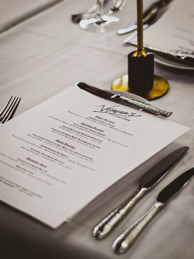 A restaurant menu sits on a table set for dinner. Silverware, a gold candle holder, and a white tablecloth are also visible.