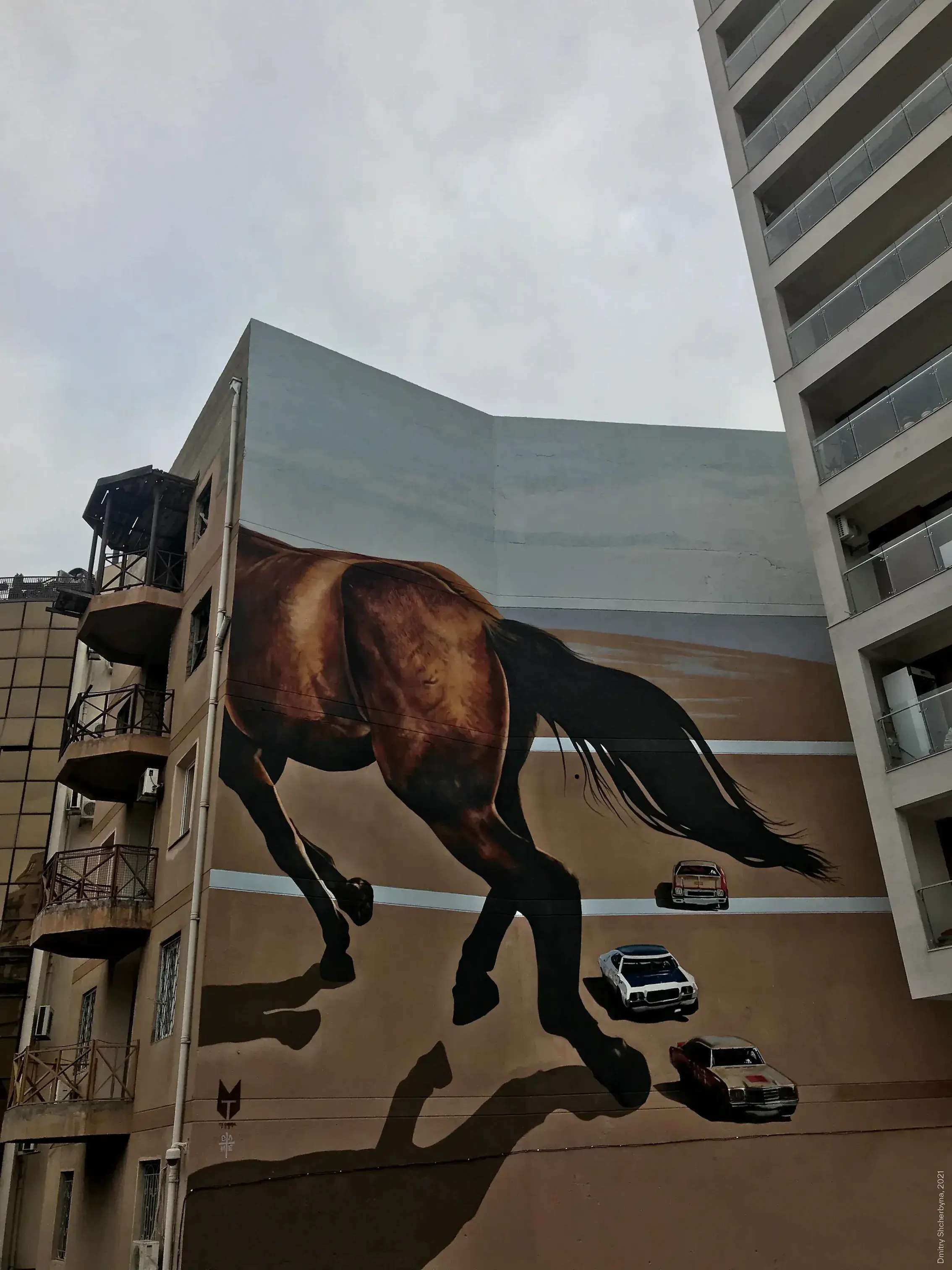 A photorealistic mural on the side of a tall building shows the rear half of a galloping brown horse, at massive scale. Three small vintage cars are on a road below it, emphasizing the dramatic size contrast.