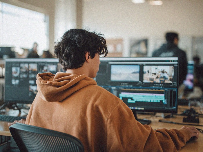 Student editing video footage on a computer with multiple monitors in a media lab.