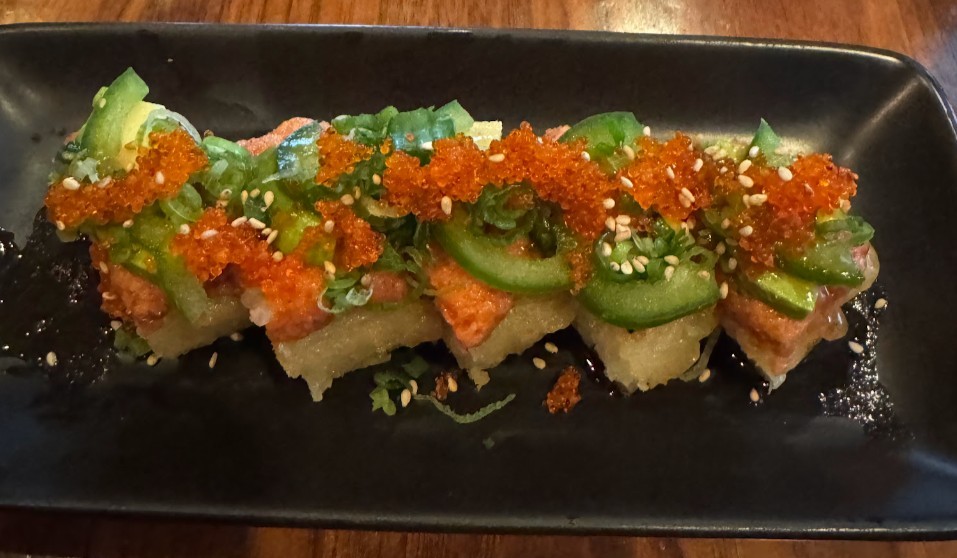 A close up of a maki sushi roll topped with red roe and jalapeno peppers and served on a black plate.