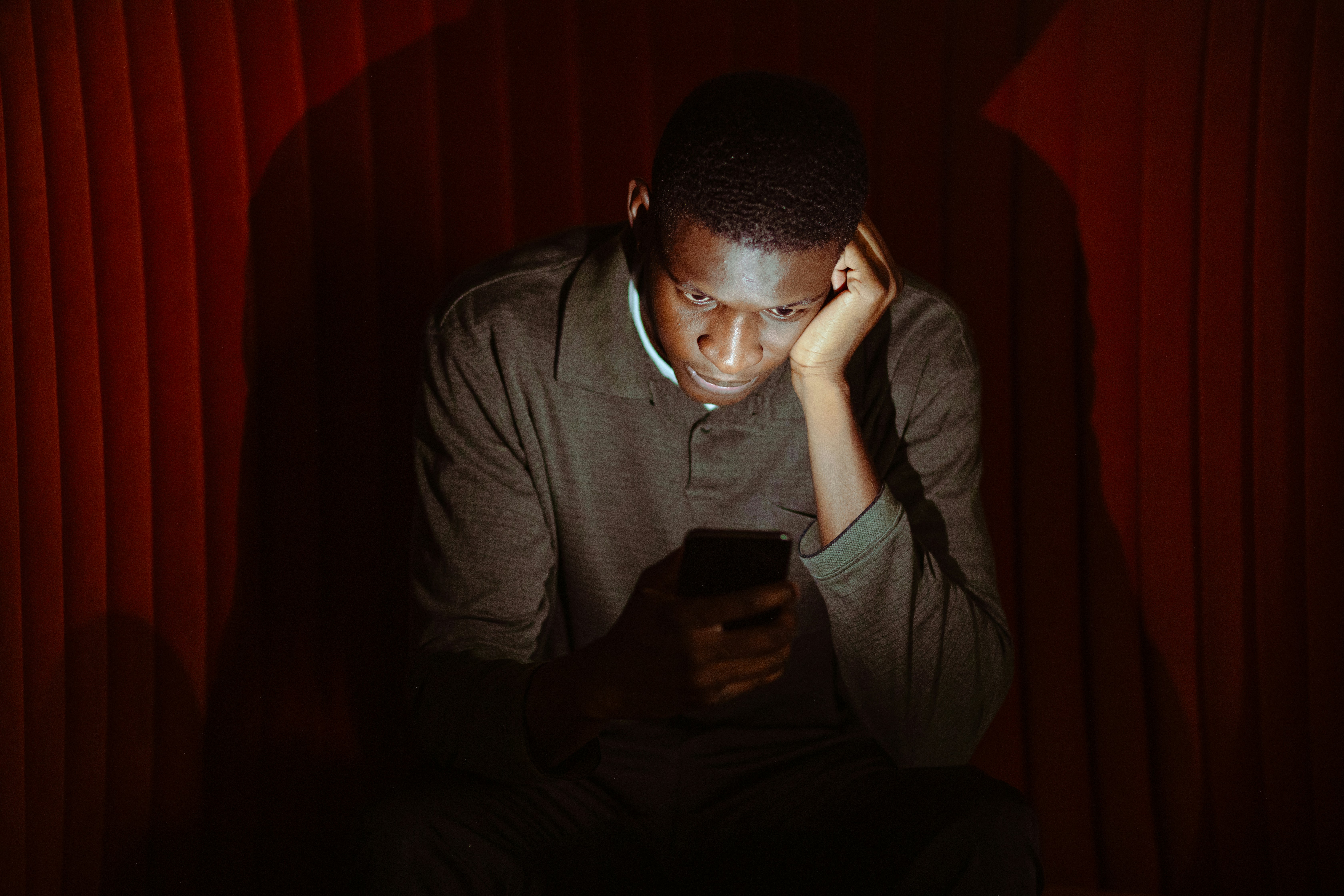 A man sitting on his smartphone in front of a red velvet curtain.