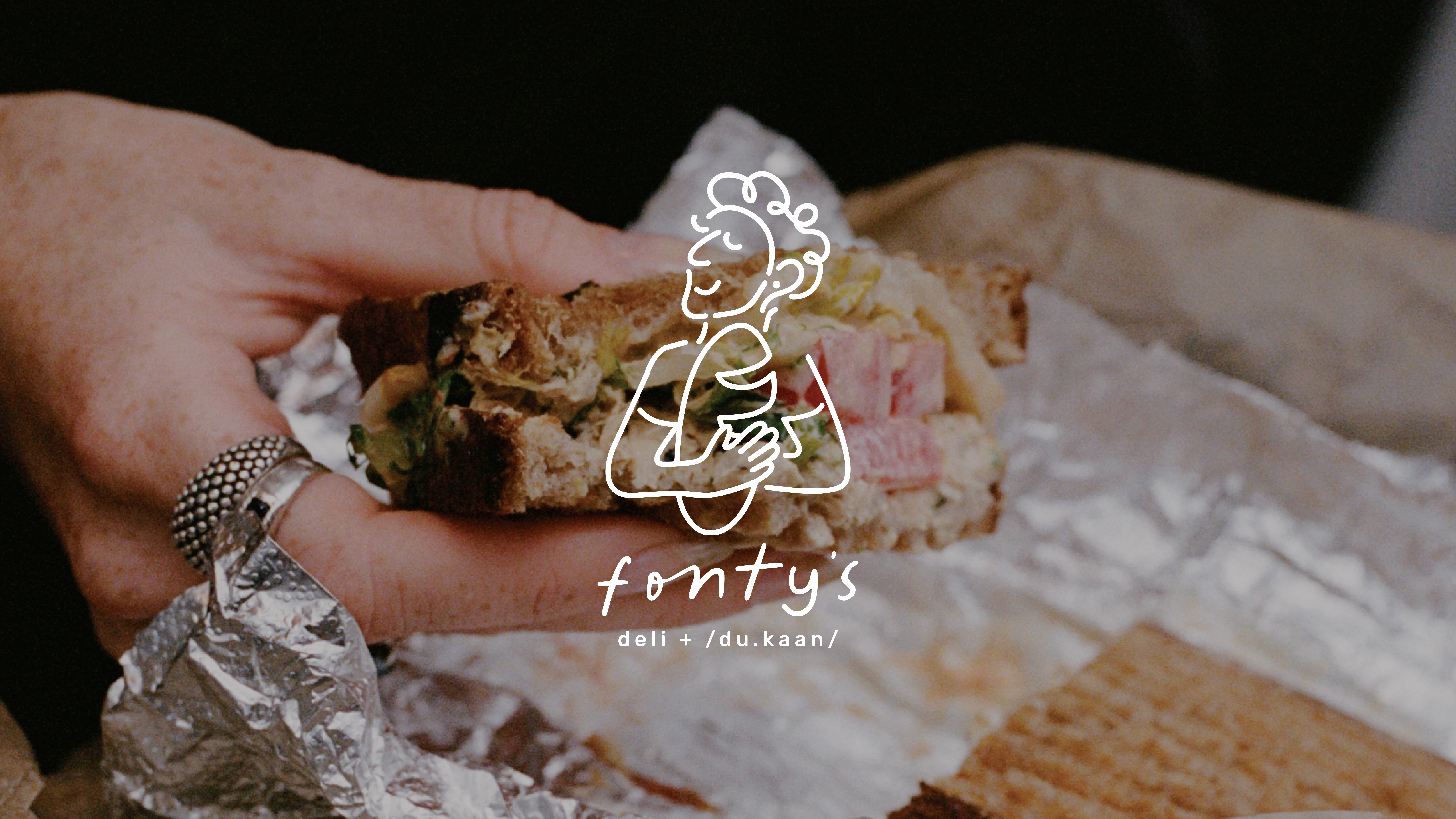 A hand holds a rustic sandwich filled with visible layers of thick-cut meat and greens, wrapped partially in foil, with a wax paper background and the logo "fontys deli" overlaid on the image.