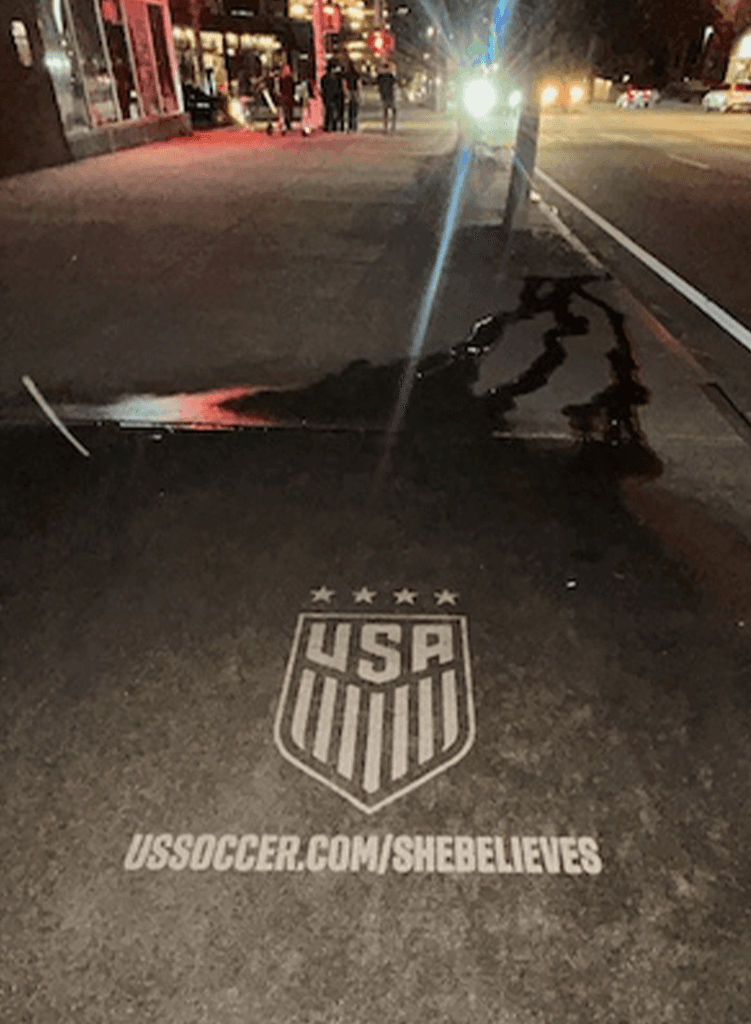 A sidewalk at night displays a USA soccer emblem, four stars, and the text ussoccer.com/shebelieves illuminated on the pavement. City lights and people are visible in the background.