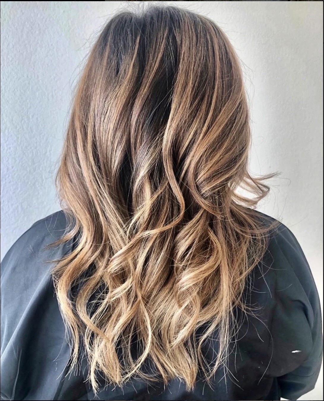 Back view of long hair with warm golden balayage and softly blended darker roots, styled in beachy waves for a sun-kissed, natural finish.