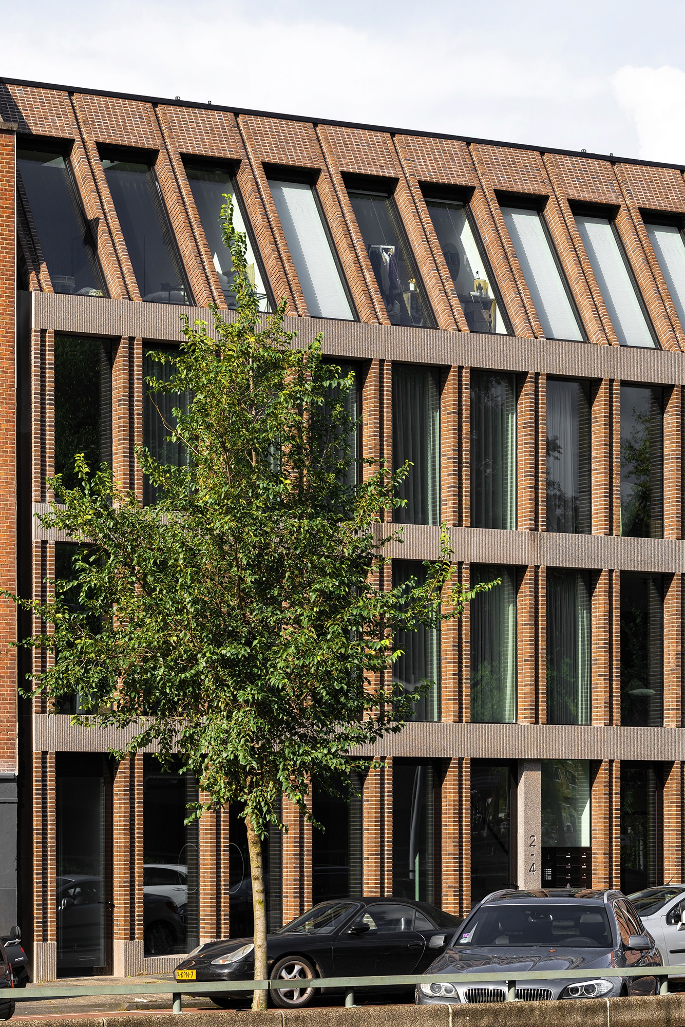 Een bakstenen gebouw met grote ramen, een boom en geparkeerde auto's ervoor.