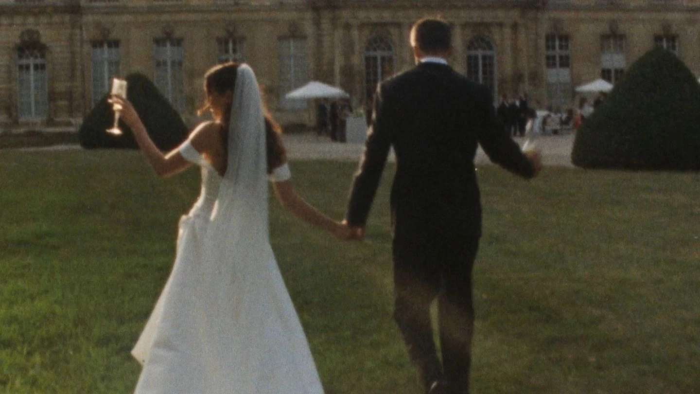 A bride and groom hold hands while walking toward a grand, historic building in a lush garden, the bride holding a champagne glass and wearing a flowing veil, capturing a romantic wedding moment.