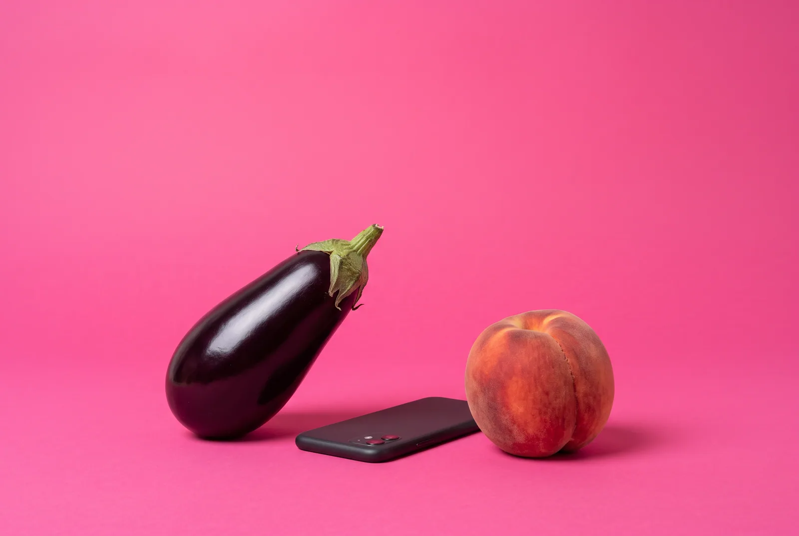 Sexting addiction concept: a glossy eggplant lying next to a smartphone placed face-down on a hot pink backdrop
