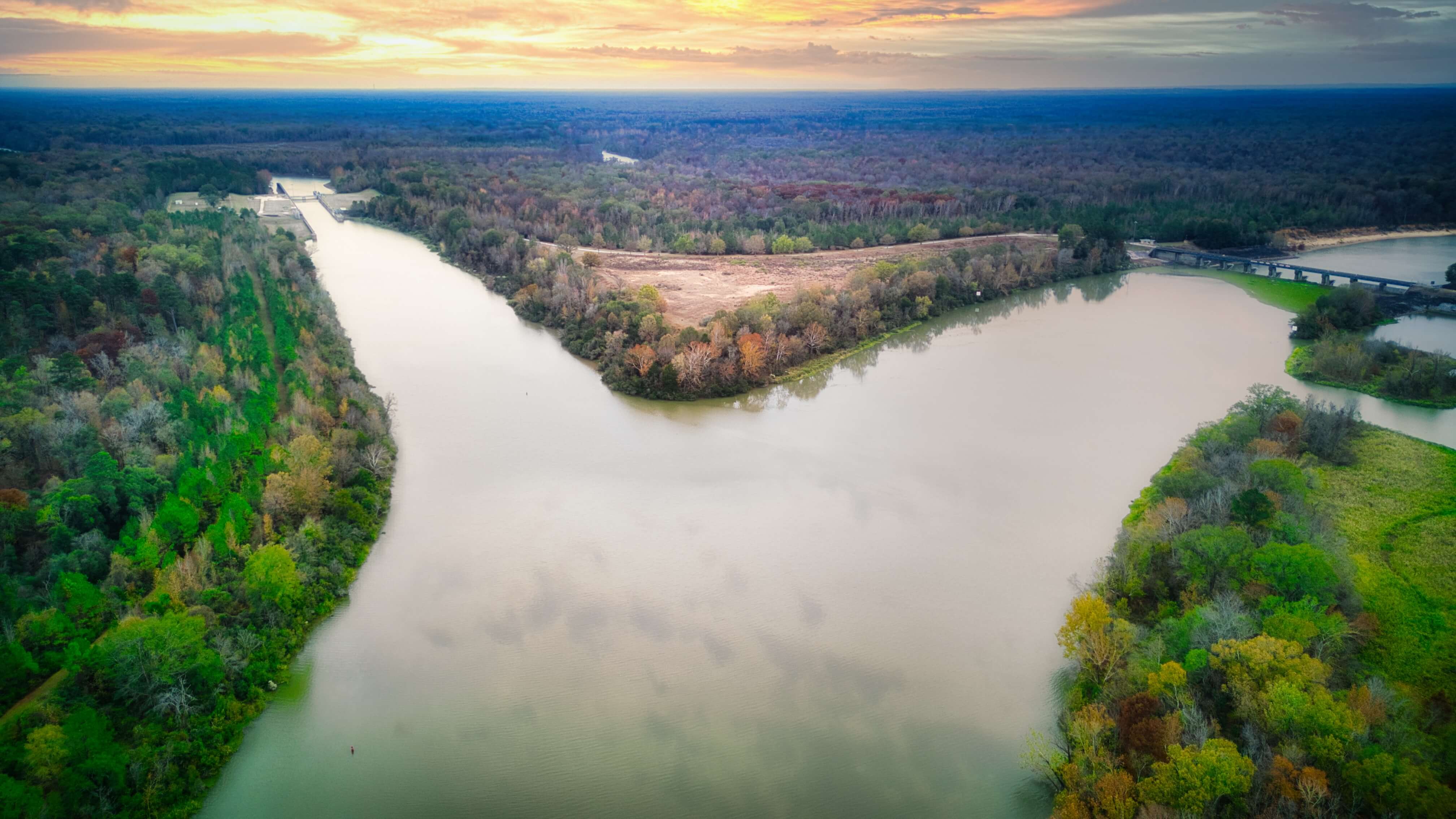 An aerial view of a wide river confluence surrounded by lush forests, with a colorful sunset sky in the background and a bridge visible in the distance