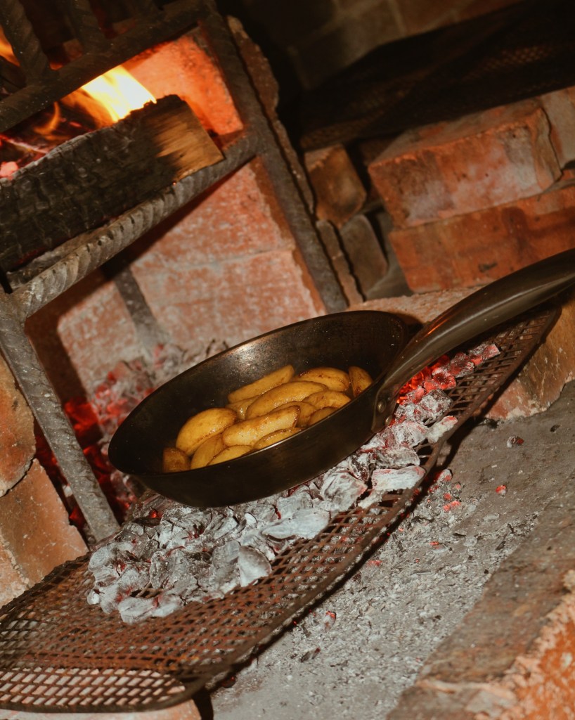 A cast iron pan with sliced apples is placed over hot coals, beside a warm fire, creating a rustic cooking scene.