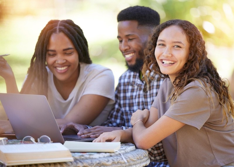 Smiling students studying together outdoors with a laptop.