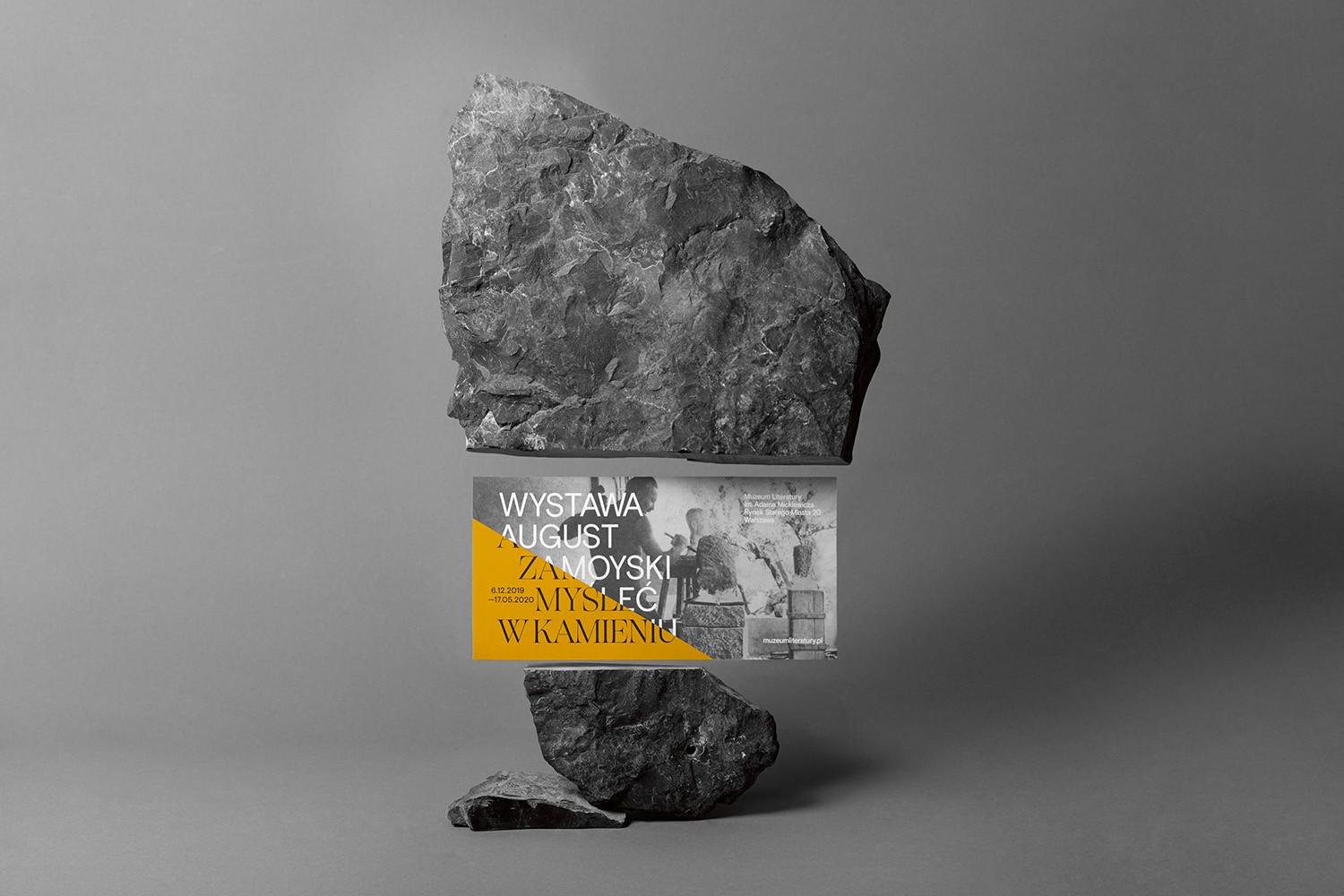 A striking visual identity mockup for the August Zamoyski exhibition, featuring a yellow and white information card physically balanced between two raw, heavy gray stones. This composition illustrates the studio's ability to create tactile brand experiences that reflect the material nature of sculpture and fine art.
