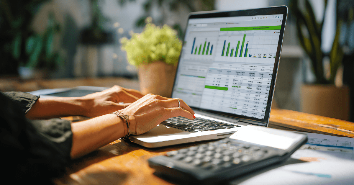 An accountant types on a laptop. There are charts and graphs on the laptop screen, and a calculator sits beside the laptop.