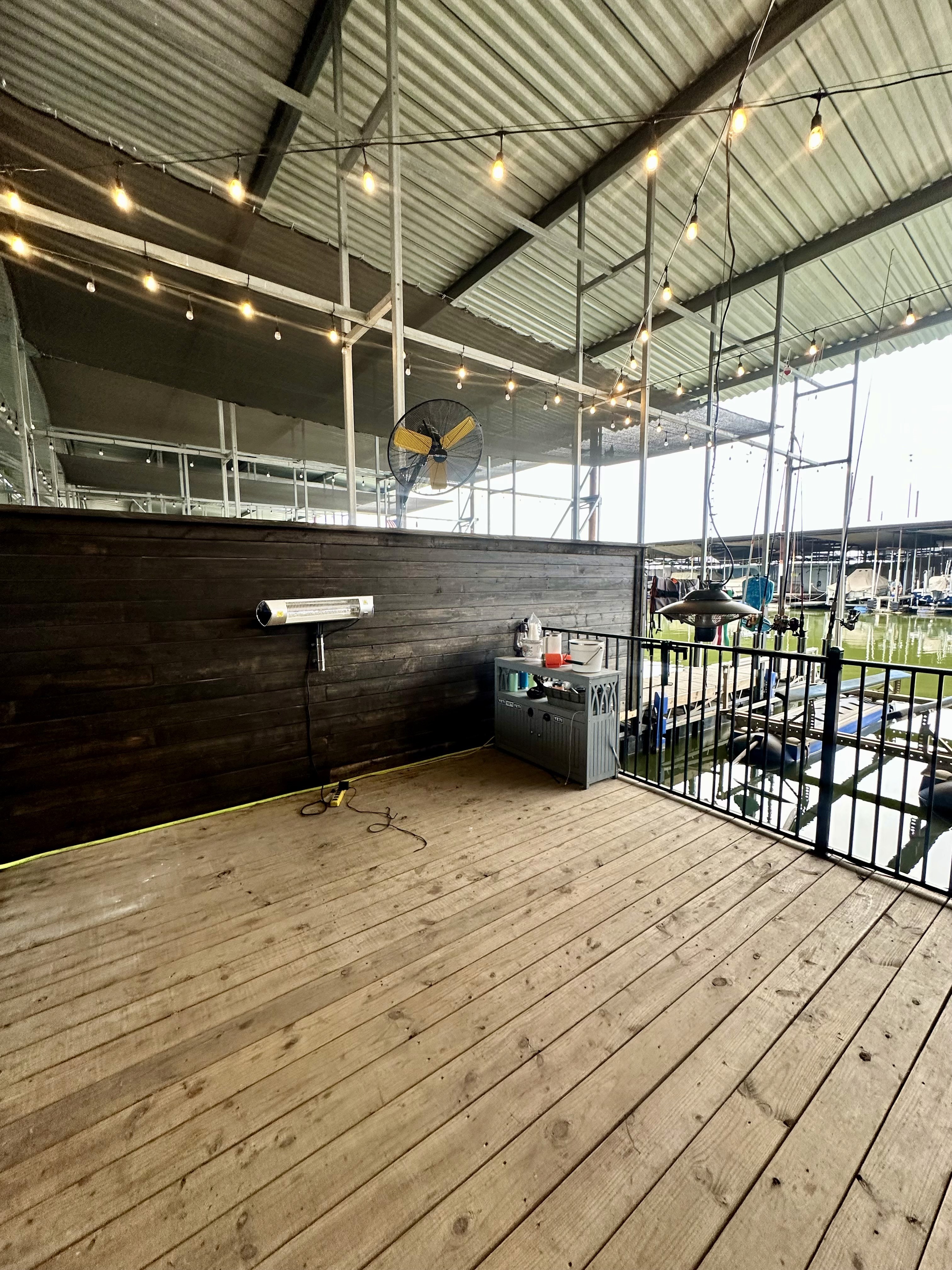 A wooden dock patio is adorned with string lights under a metal roof, overlooking a marina with boats, and features a wall-mounted heater and a utility sink.