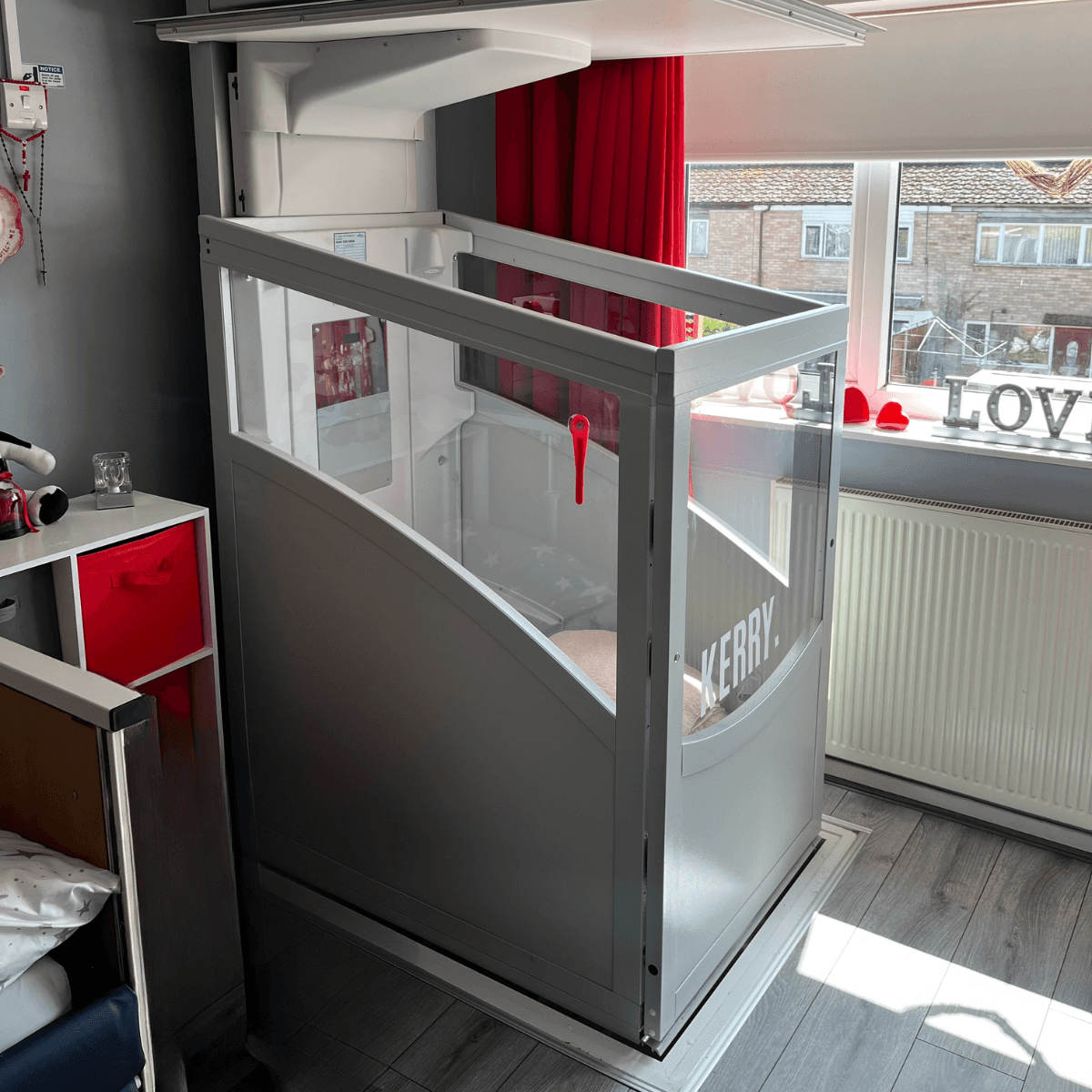 through-floor home lift installed in a UK residential bedroom — grey aluminium frame with toughened glass panels, red emergency handle visible, ceiling aperture showing upper floor level, compact domestic installation on timber flooring
