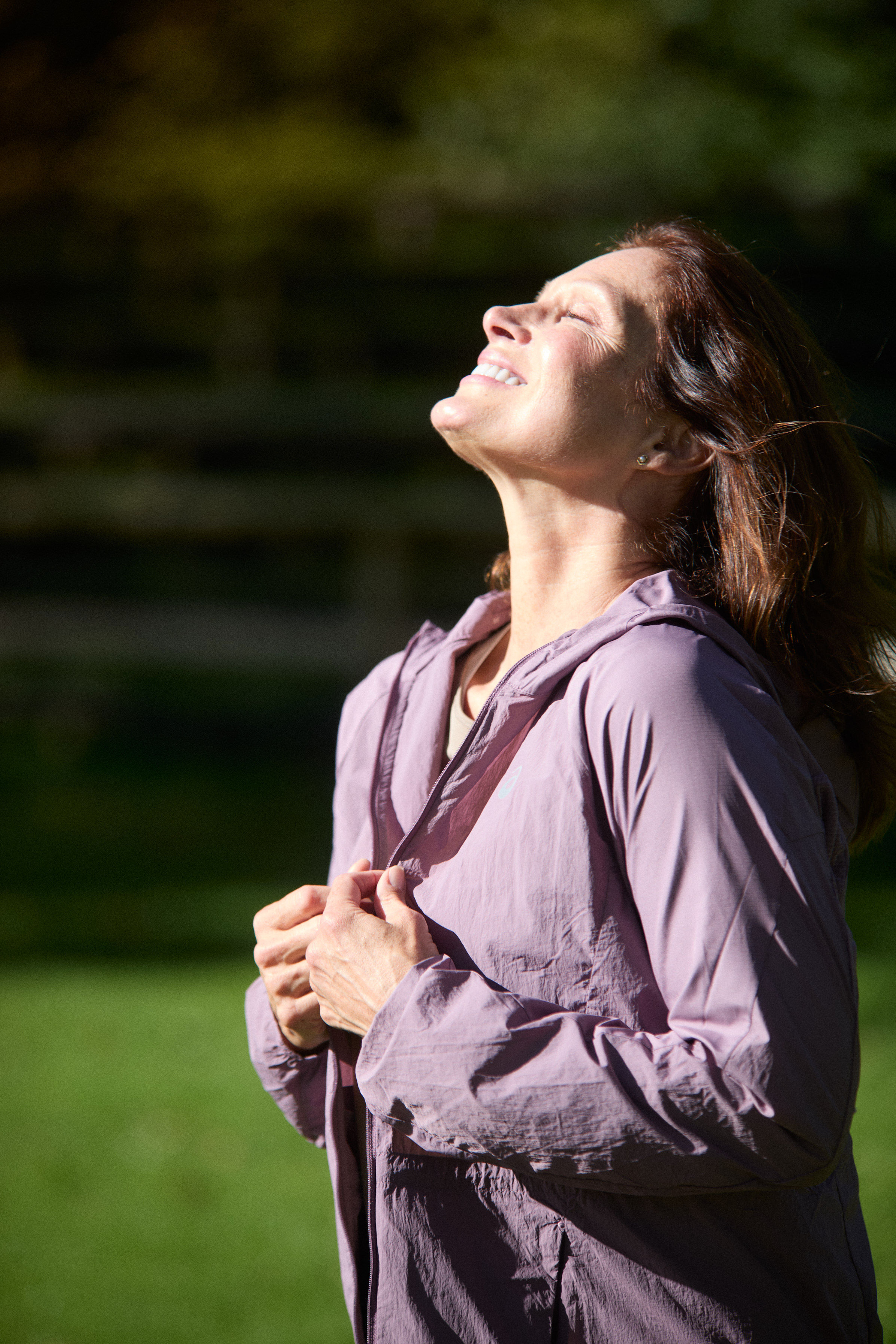 Woman sitting in sunlight with eyes closed, wearing relaxed neutral clothing in a peaceful setting.