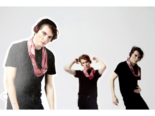Three young men stand against a plain light gray background. The man on the left wears a black t-shirt and a pink scarf around his neck, and red over-ear headphones. The man in the center has his hands raised near his head. The man on the right wears a black t-shirt and a pink scarf. All three are looking directly at the camera.