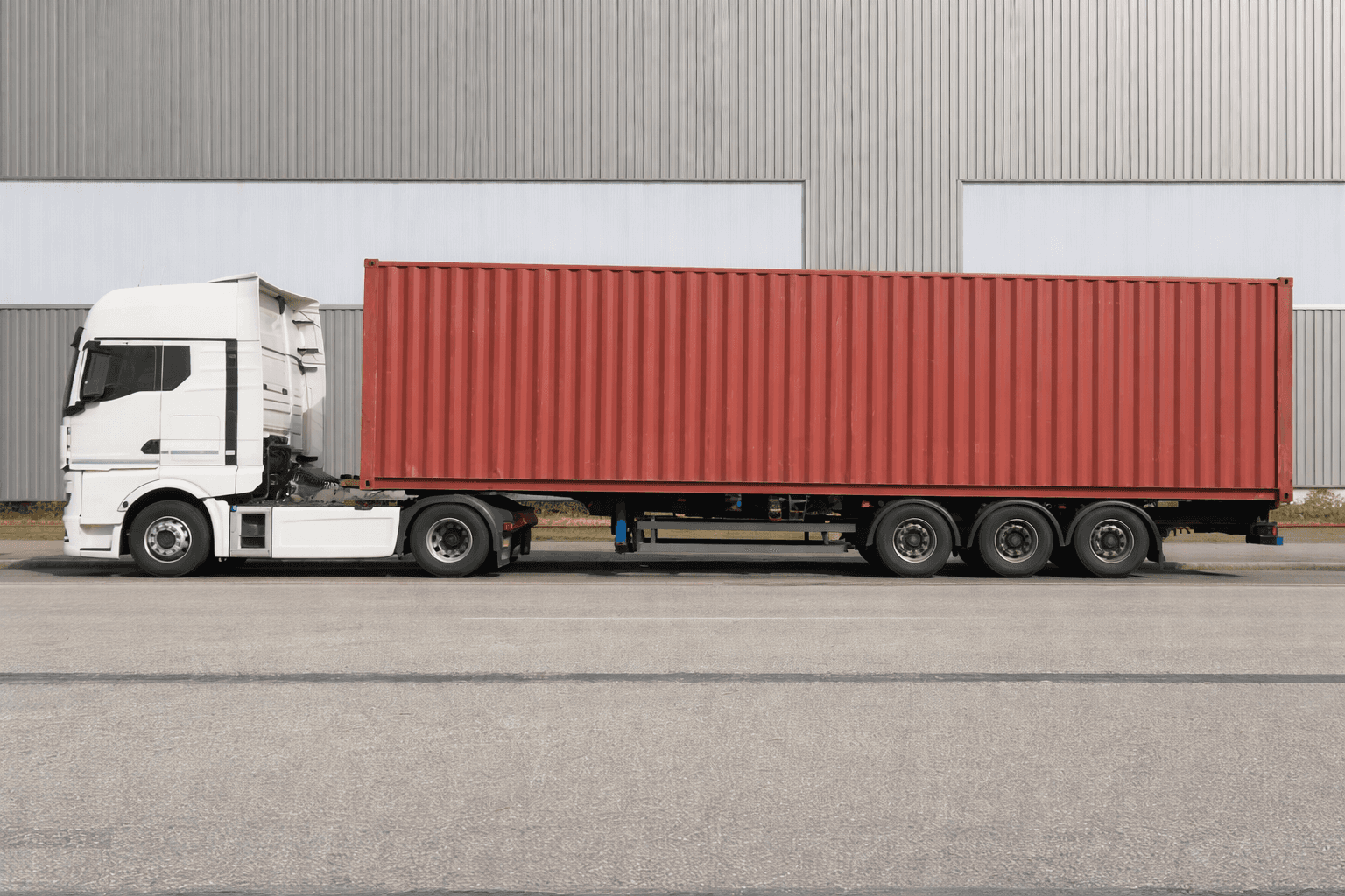 White semi-truck with a red shipping container parked beside a warehouse.