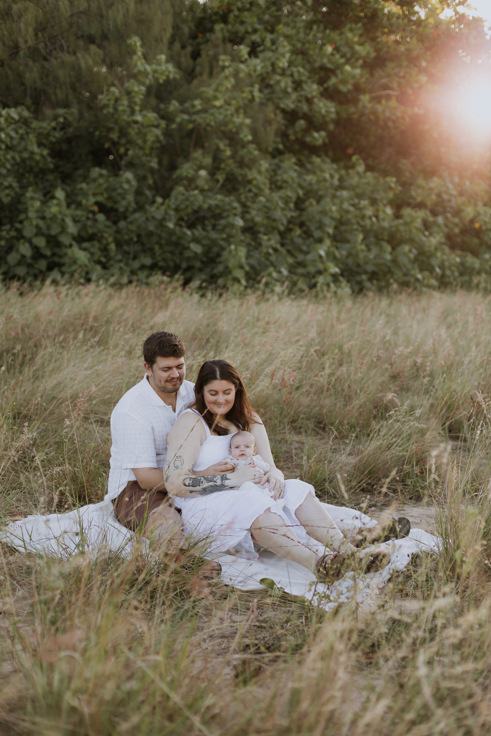 Parents sitting in the long grass holding newborn baby with glowing sun in Mackay
