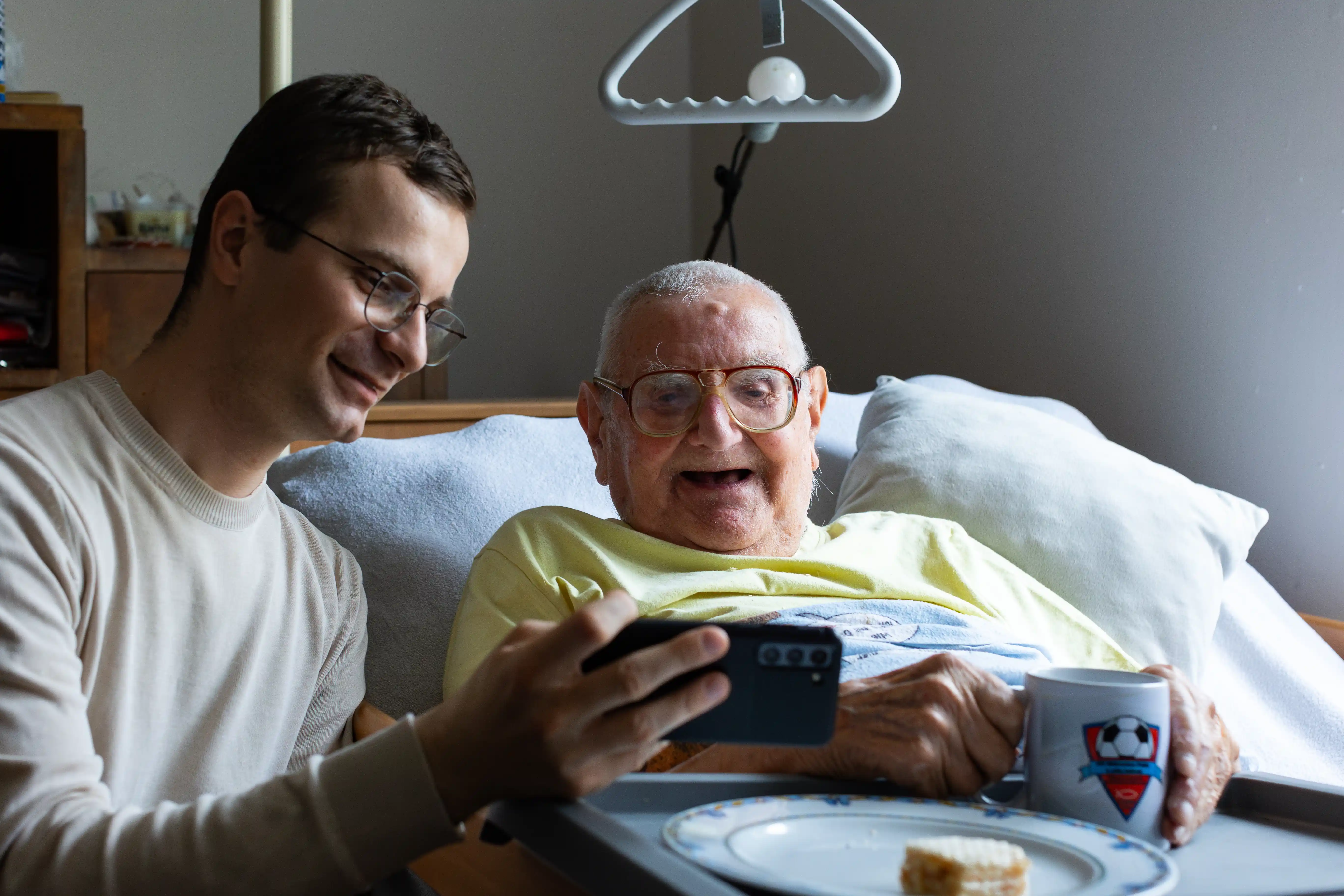 Młody mężczyzna pokazuje coś na swoim telefonie starszemu mężczyźnie leżącemu w łóżku, który trzyma w ręku kubek. Nad nim wisi chwytak; na jego kolanach znajduje się taca. Oboje się uśmiechają.