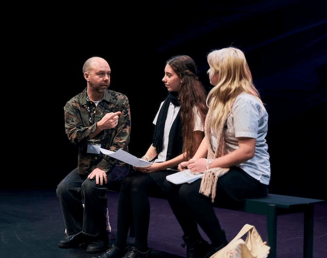 A man wearing a camouflage jacket gestures while speaking to two teenage girls seated on a bench with scripts in hand during a drama workshop on a dark theatre stage.