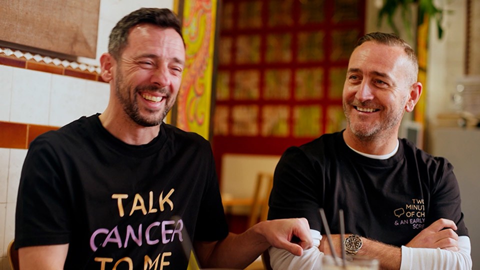 Two men, Ralph Little and Will Mellor,  laughing together at a café table.