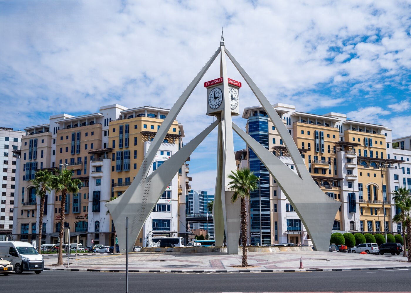 Iconic Deira Clocktower in Dubai roundabout, framed by palm trees, traffic, and mid-rise buildings under a partly cloudy sky.