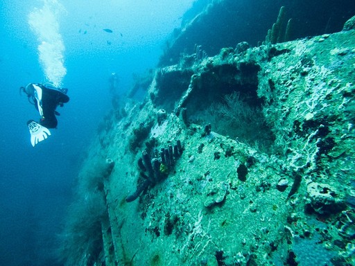 Scuba diver exploring a large underwater shipwreck covered in coral and marine life in clear blue water.