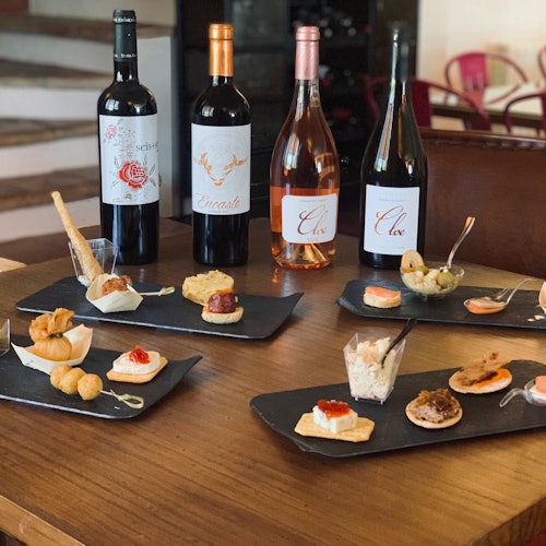 Valikoima alkupaloja puisella pöydällä, taustalla neljä viinipulloa, viihtyisässä sisätilassa.