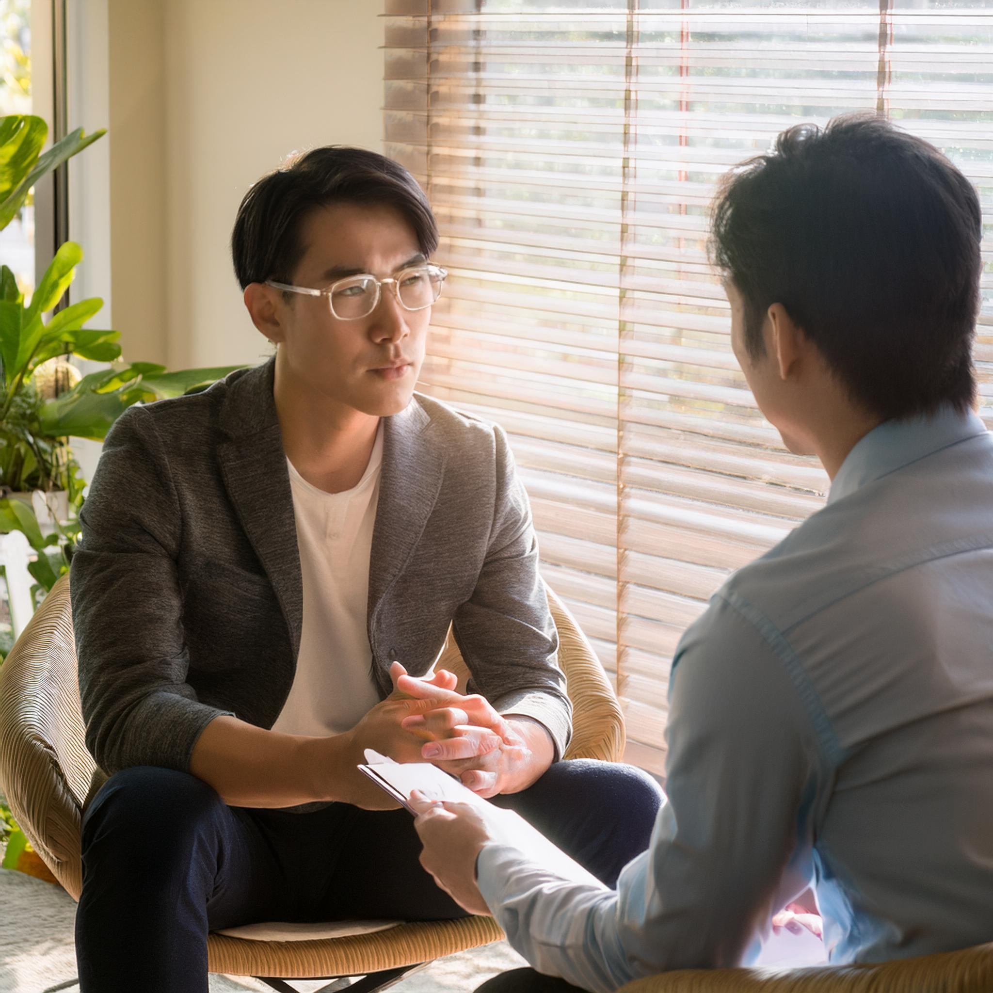 Two men talking by a window, one holding a clipboard and the other wearing glasses.