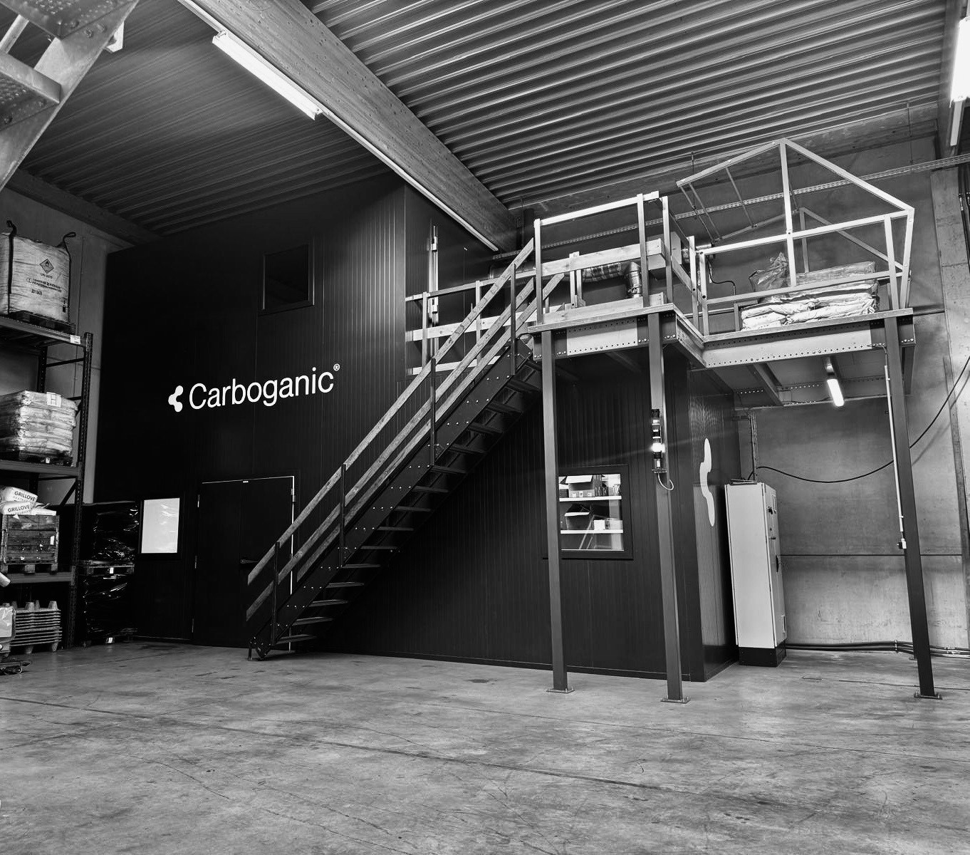 A black and white wide shot of an industrial interior featuring a large, dark-paneled production unit with the "Carboganic" logo. A metal staircase leads to an elevated mezzanine platform, and heavy-duty storage racks with large industrial bags are visible to the left.