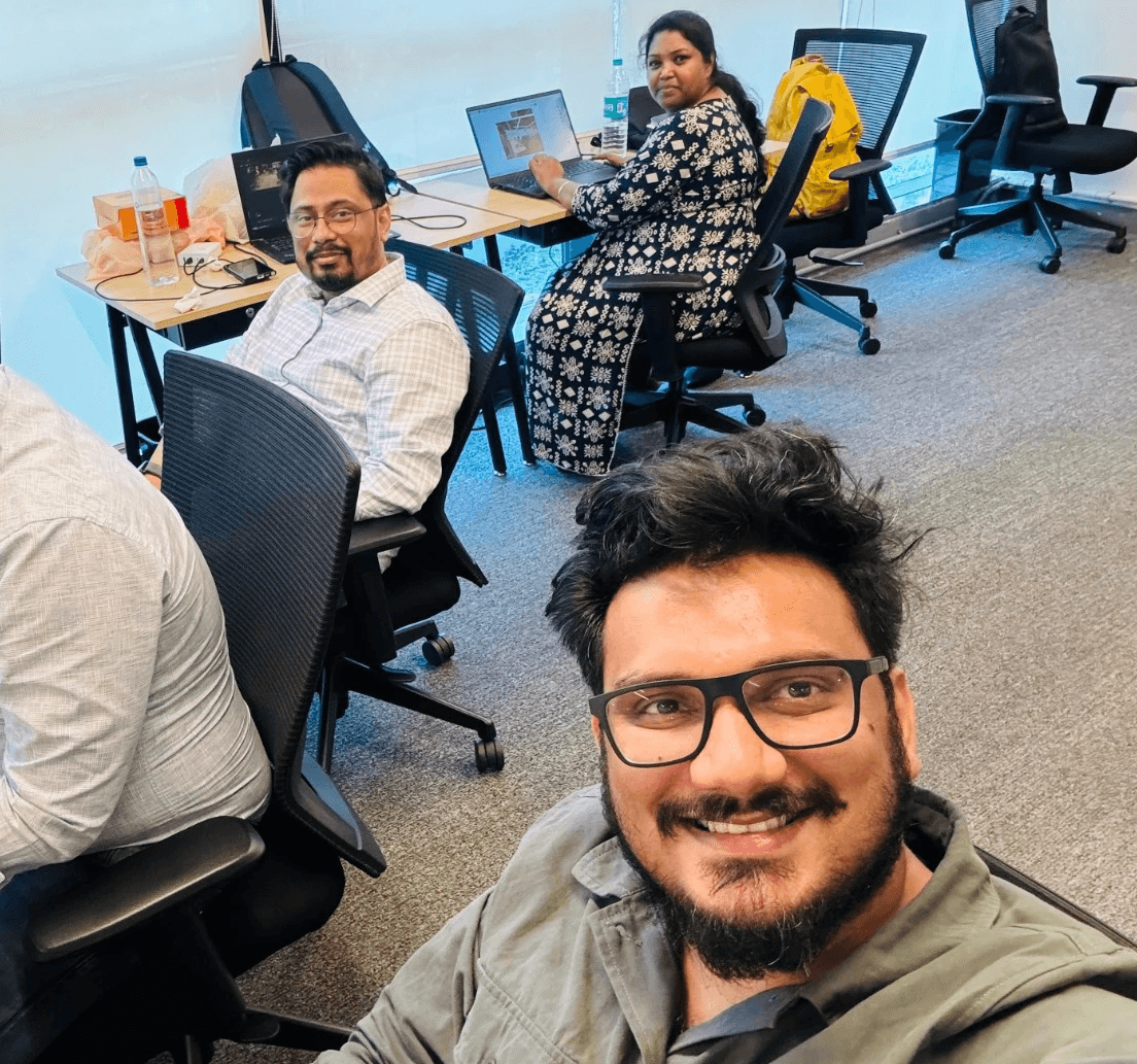 A group of people working in an office, with one person smiling for a selfie in the foreground.