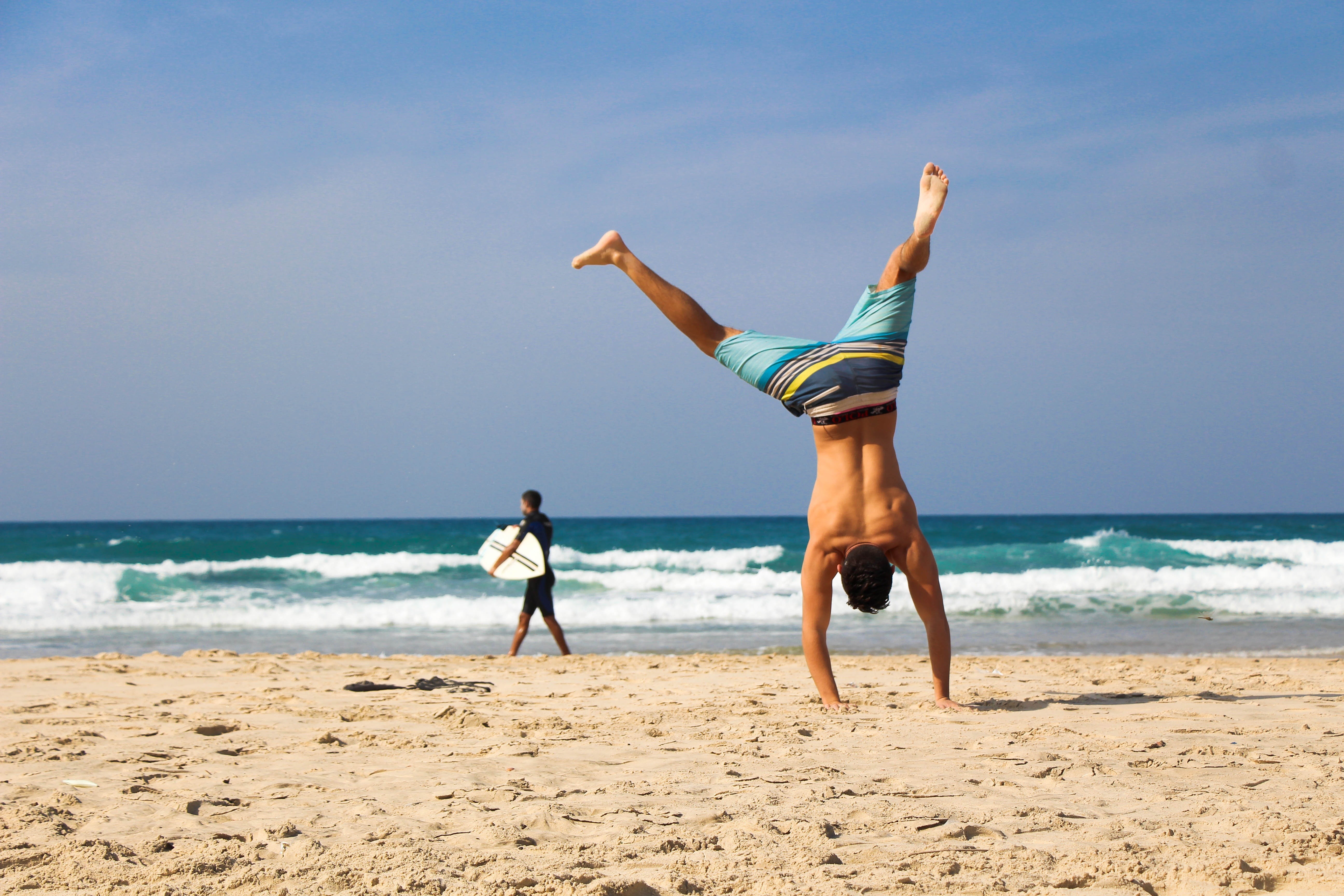 Man on a beach doing a handstand with another man holding a surfboard
