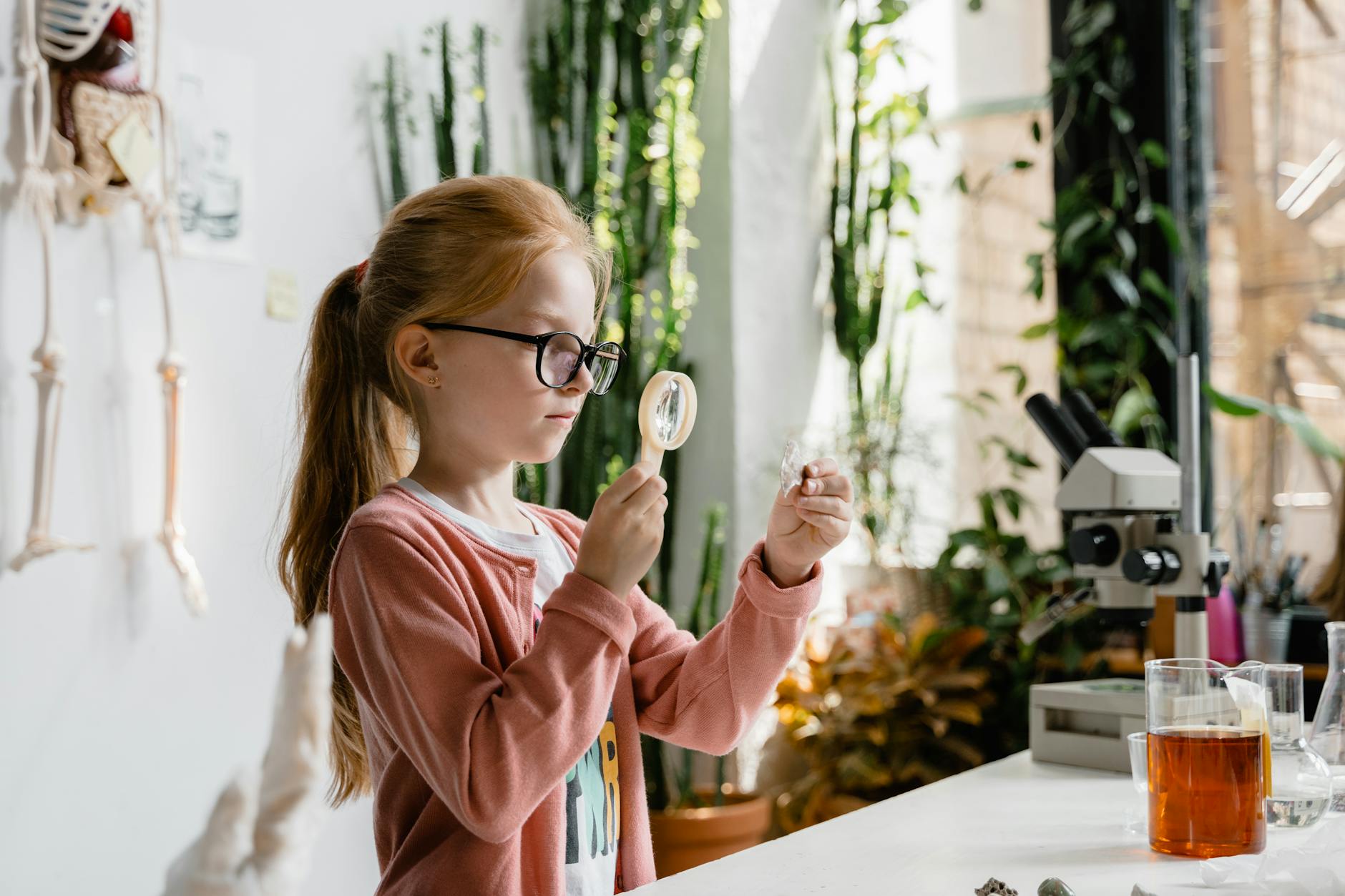 A curious young student looks through a magnifying glass at a green leaf in a sunny classroom.