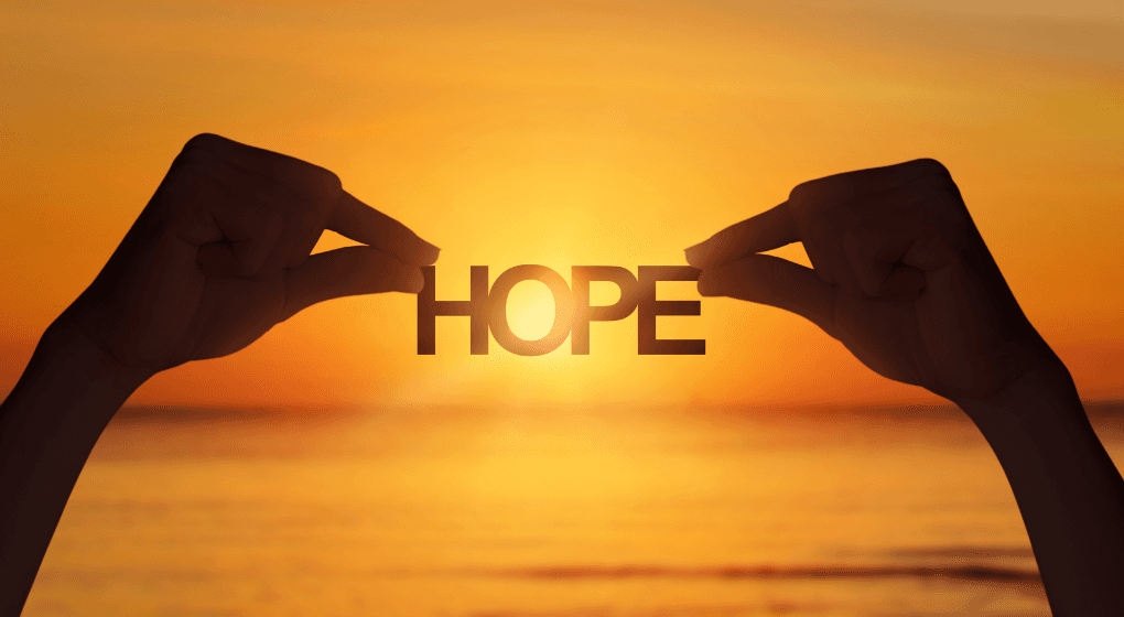 Image of two hands holding a sign that says "hope" with the sun setting behind the ocean behind the word.