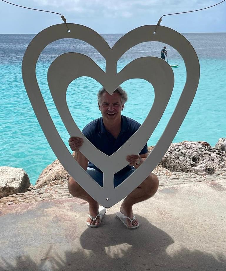 Smiling female therapist with short gray hair, wearing a brown top and a purple shawl, surrounded by a cozy background.