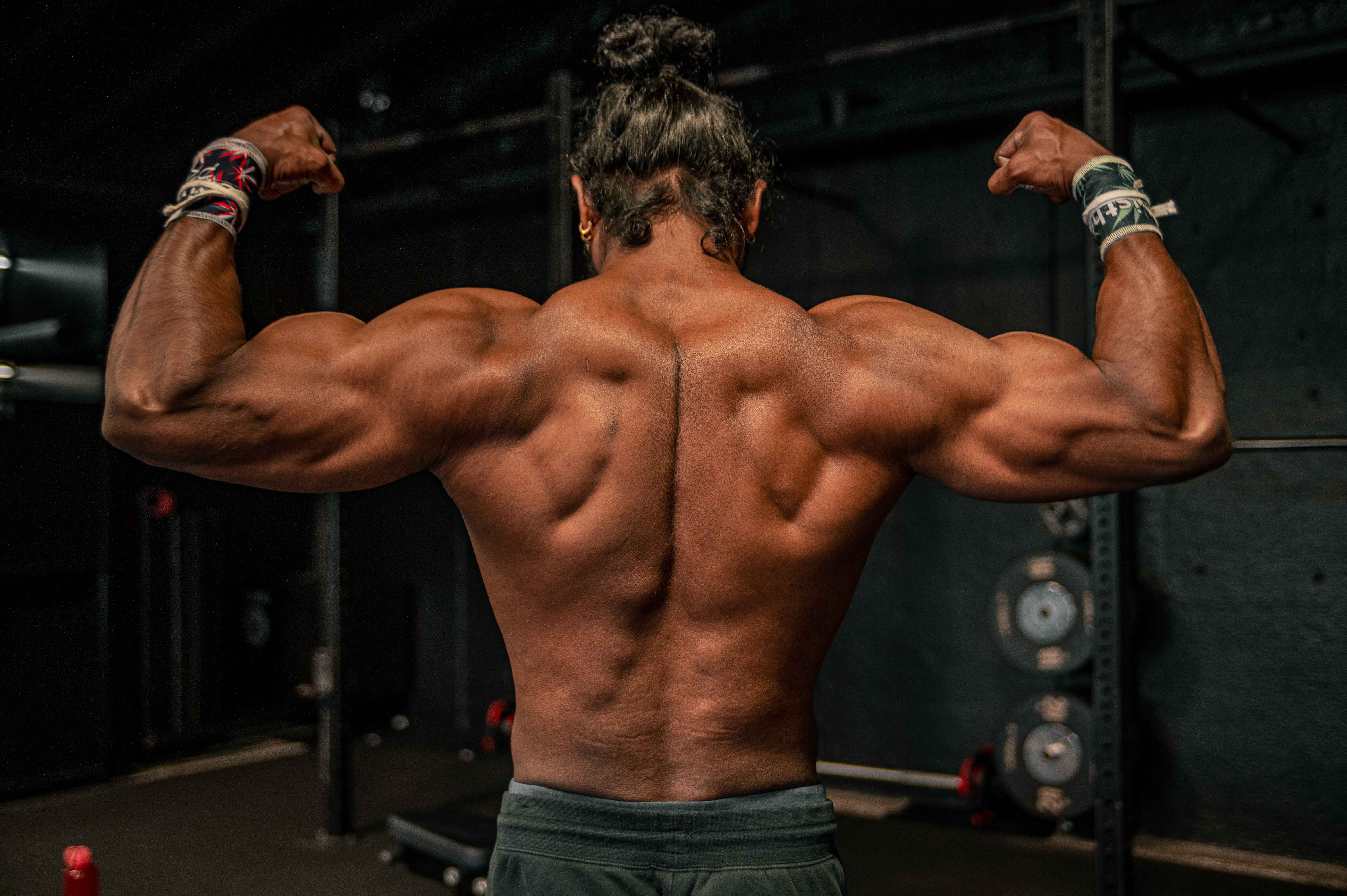 A muscular bodybuilder with long hair flexes his back and biceps in a gym, showcasing defined muscles.