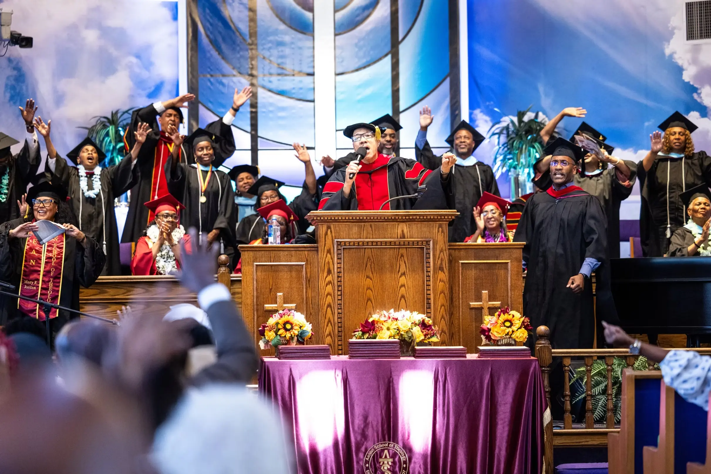 Graduates in caps and gowns celebrate on stage at the Aenon Bible College 2026 Graduation ceremony, with arms raised in joy beneath a decorated sanctuary altar adorned with floral arrangements.