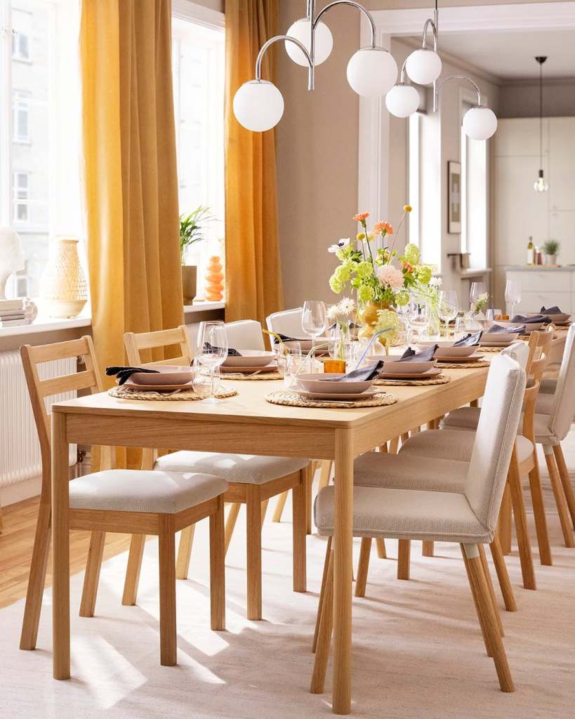 A beautifully set dining table with natural wood finish, surrounded by light-colored chairs. The table is adorned with stylish tableware, glasses, and a decorative floral arrangement, while warm curtains frame the bright windows.