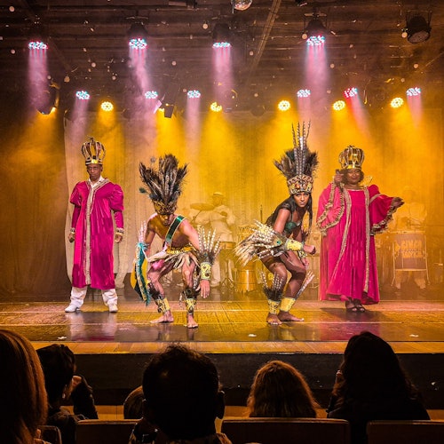 四位表演者身着传统且华丽的服装在舞台上表演，前景中是正在观赏的观众。