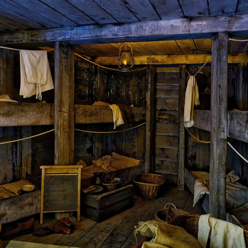 Dimly lit, rustic cabin with wooden bunk beds, hanging laundry, a lantern, a washboard, and storage baskets.