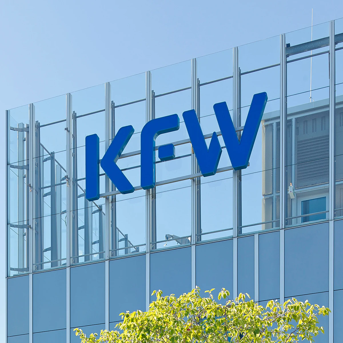 Glasfassade eines Gebäudes mit einem großen blauen „KFW“-Logo und grüner Bepflanzung im Vordergrund.
