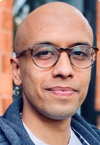 a bald man with brown skin and glasses looking into camera