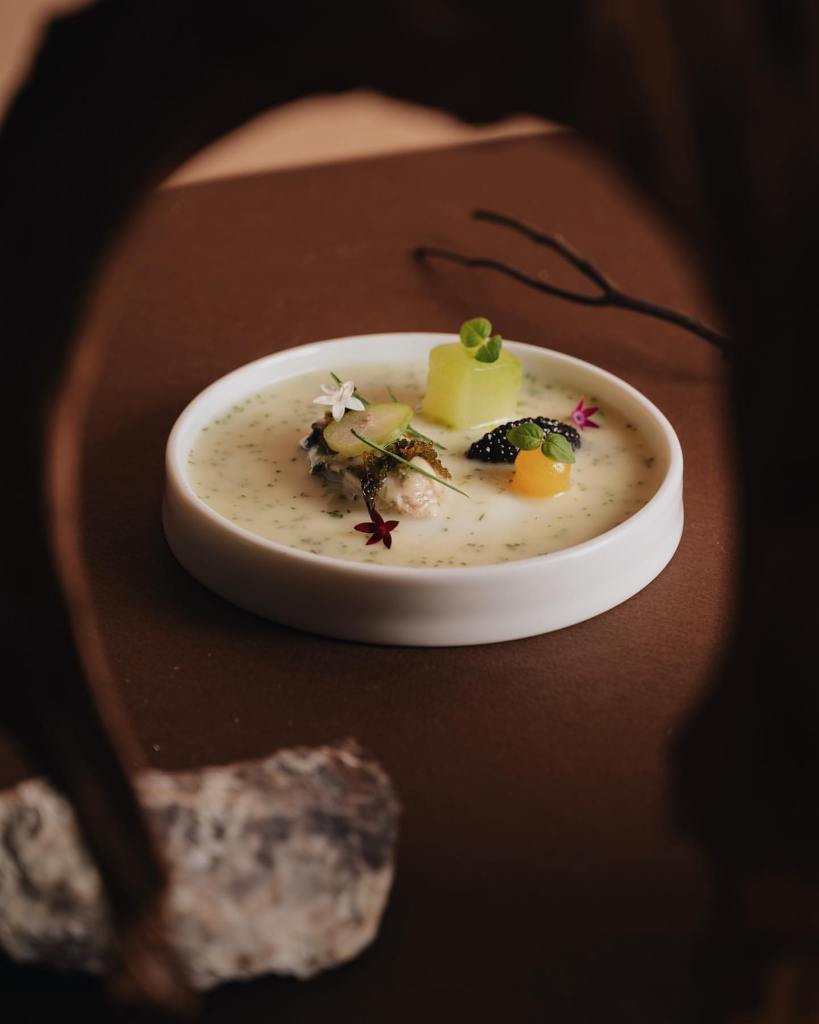 A beautifully plated dish featuring caviar, garnished with herbs, colorful vegetables, and a creamy sauce, presented in a white bowl on a brown surface.