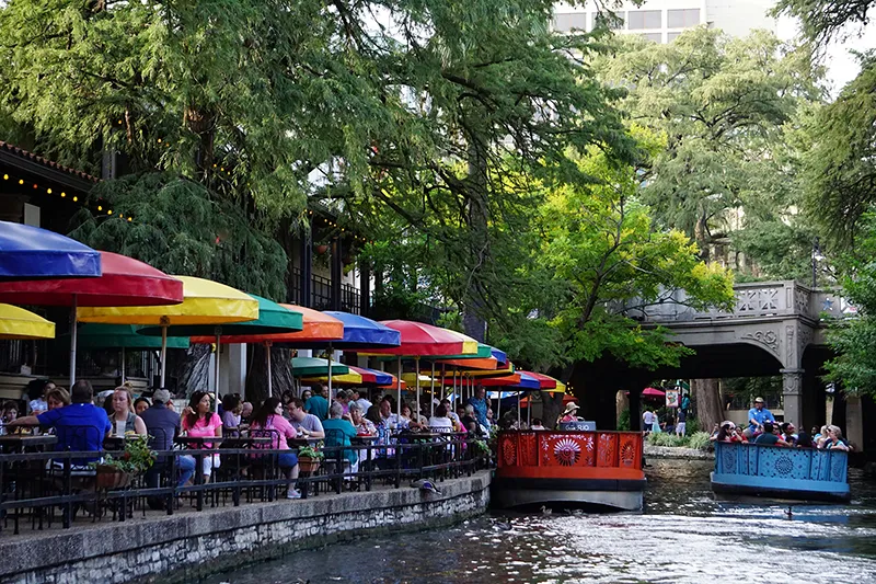 Riverside patio dining under colorful umbrellas along the San Antonio River Walk, with river barges passing beneath a stone footbridge