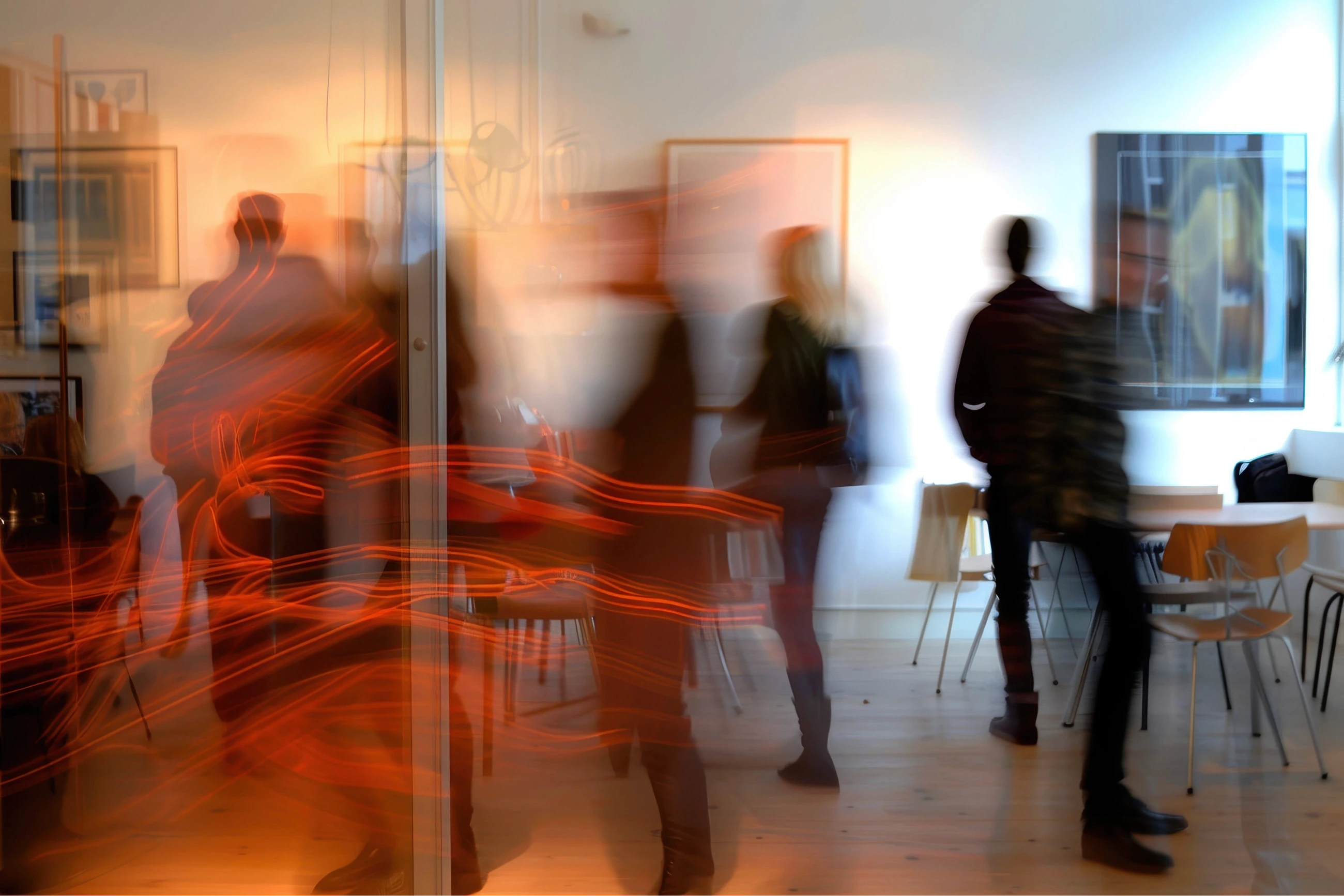 People are captured in motion, creating a dynamic blur effect, within a modern, art-filled room with wooden flooring and minimalistic furniture.